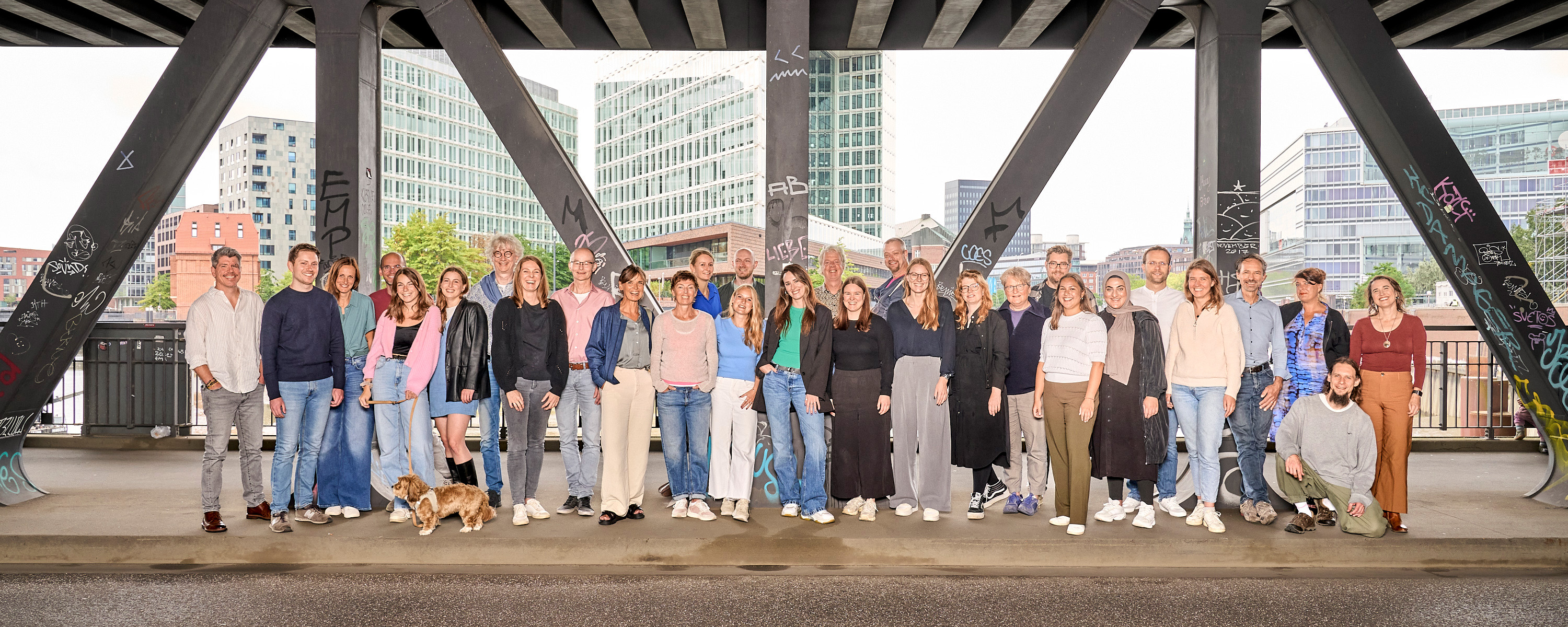 Gruppenbild mit bunt gekleideten Menschen auf einer Brücke vor städtischem HIntergrund
