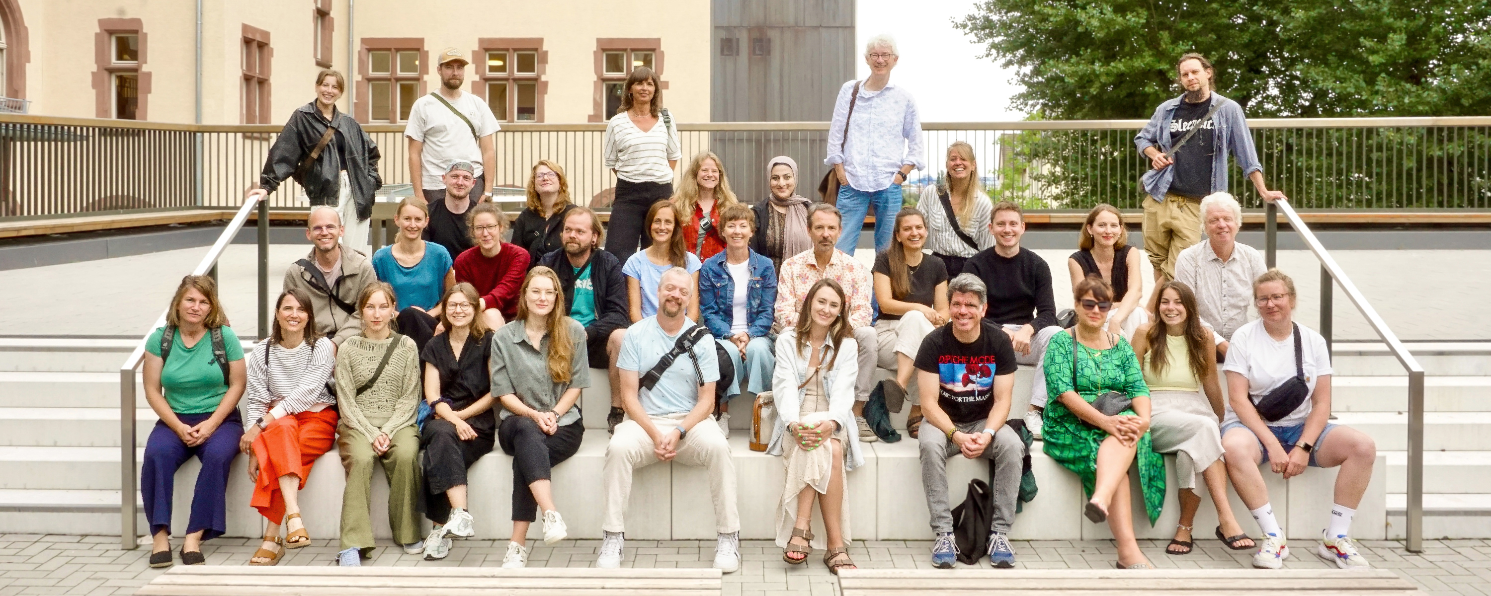 Eine Gruppe von erwachsenen Menschen sitzt und steht auf einer Sitzstufenanlage eines Schulhofs.