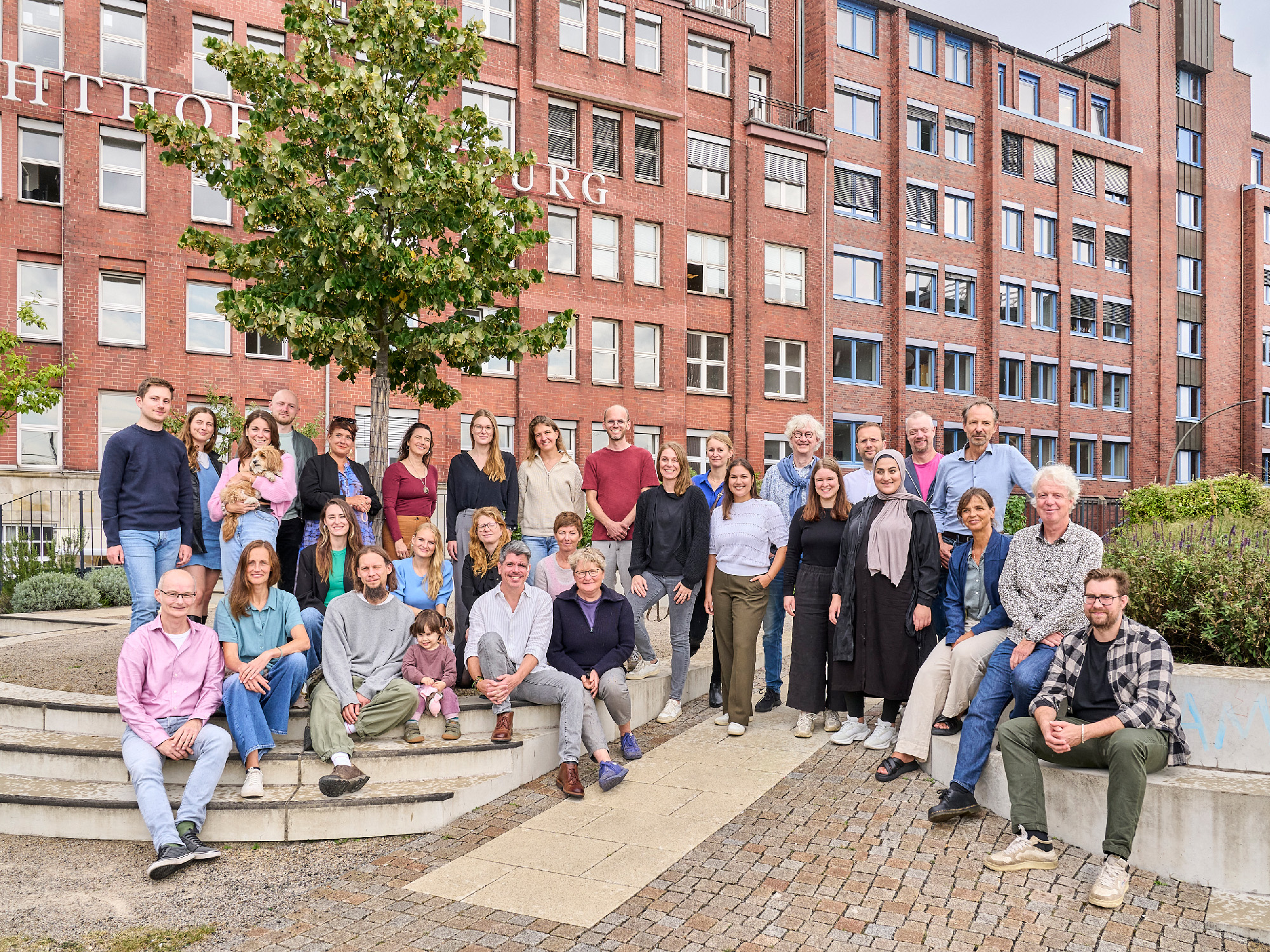 Gruppenbild mit bunt gekleideten Menschen auf einem gestalteten Platz vor großem Gebäude aus Backstein