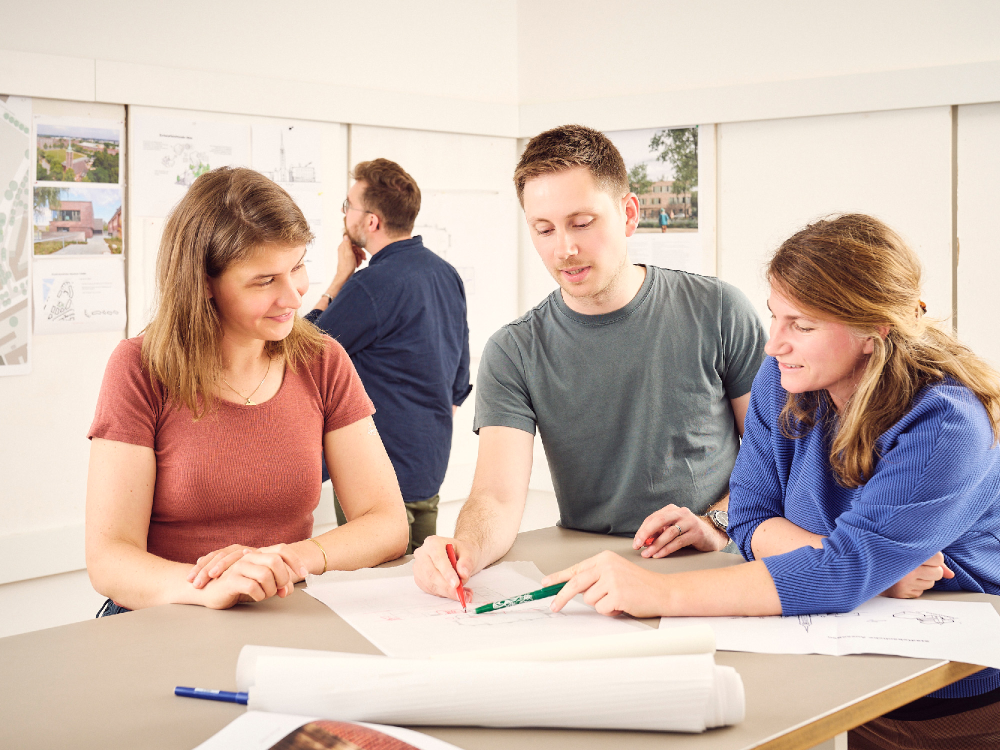 Eine kleine Gruppe Menschen steht um einen hohen Tisch herum, auf der einer Architekturzeichnung liegt, und diskutiert.