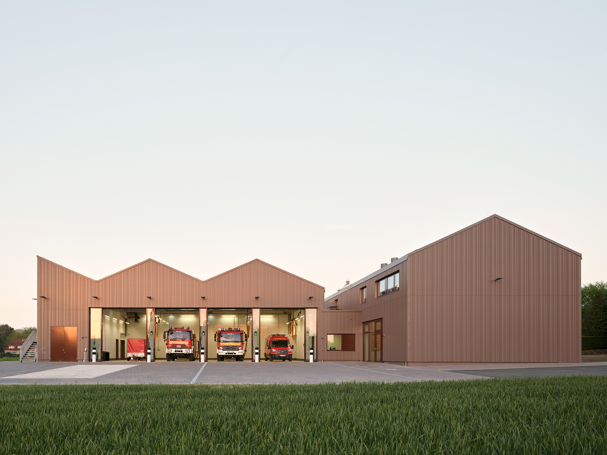 Eine Außenaufnahme eines Feuerwehrhauses mit geöffneten Toren zur Fahrzeughalle, in der vier Einsatzfahrzeuge stehen. Die Fassade ist aus rotbraunem Metall.