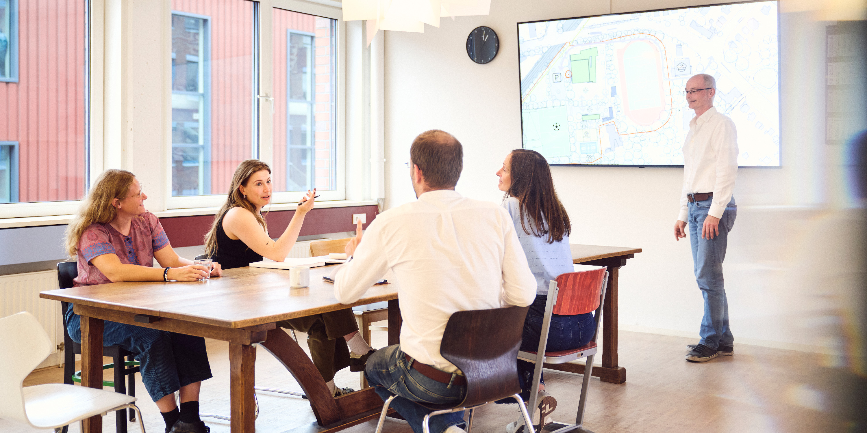 Ein Mann steht vor einem großen an der Wand befestigten Bildschirm und präsentiert etwas. Vier Architekten sitzen im Vordergrund an einem langen Tisch und diskutiert.