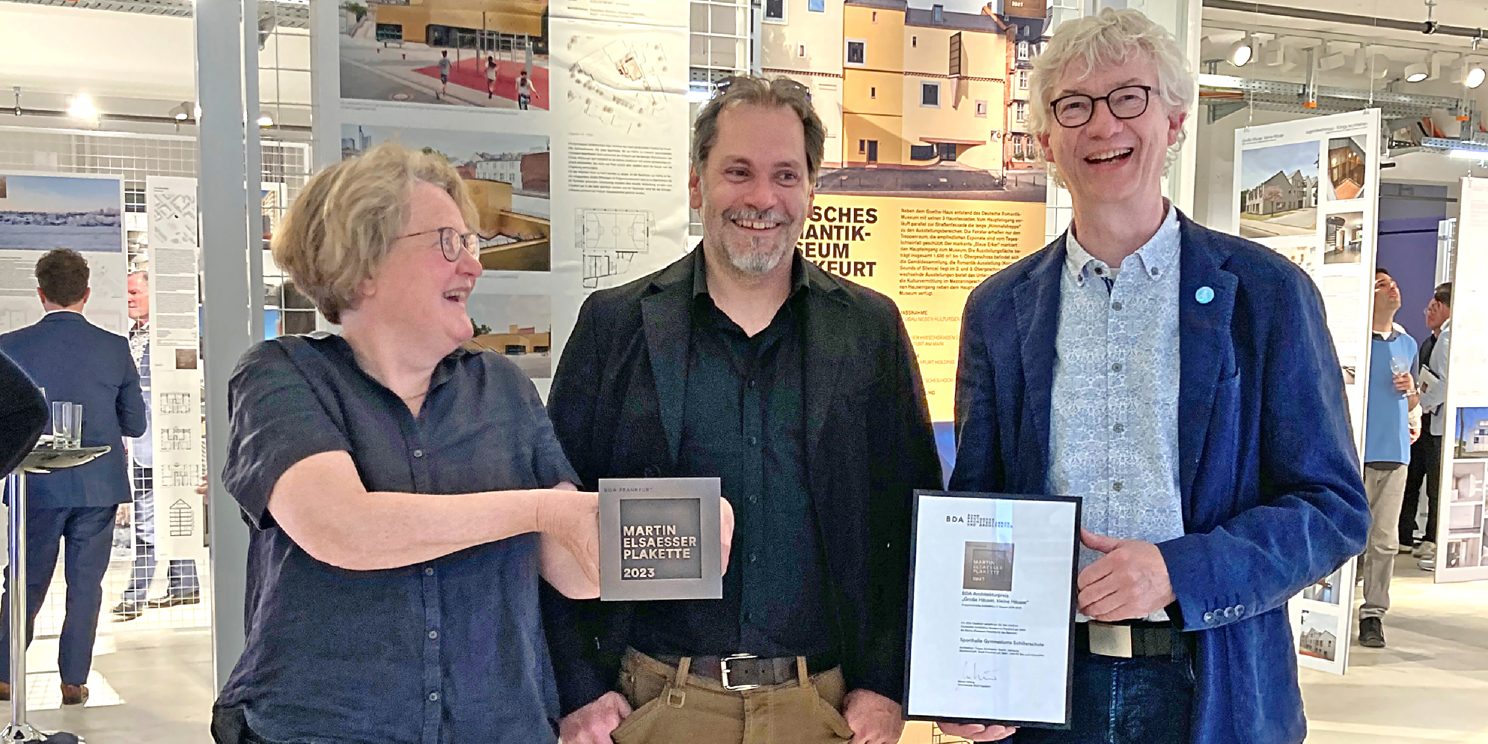 Eine Frau und zwei Männer in einer Ausstellung. Sie zeigen Architektur Auszeichnungen in Form einer Plakette und einer Urkunde.