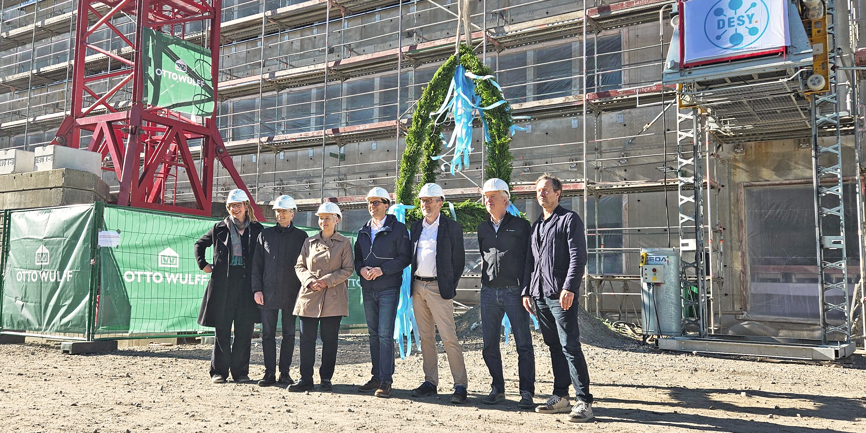 Eine Gruppe von Menschen mit weißen Bauhelmen stehen vor einem Rohbau mit einem geschmückten Richtkranz