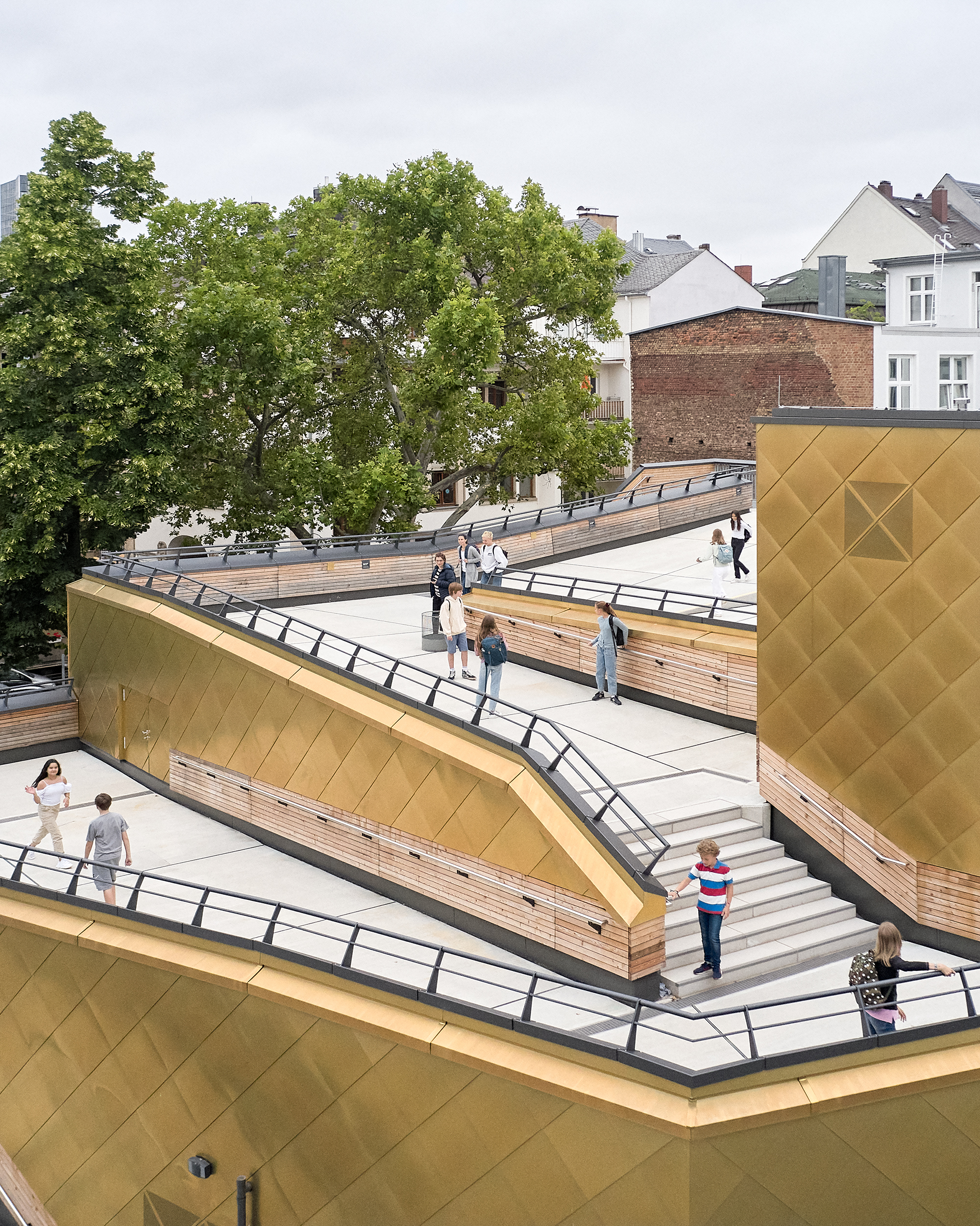 Schüler verteilen sich über Treppen und Rampen auf dem begehbaren Dach einer mehrfach gestaffelten Sporthalle. Die Fassade besteht aus goldenen Metalschindeln.