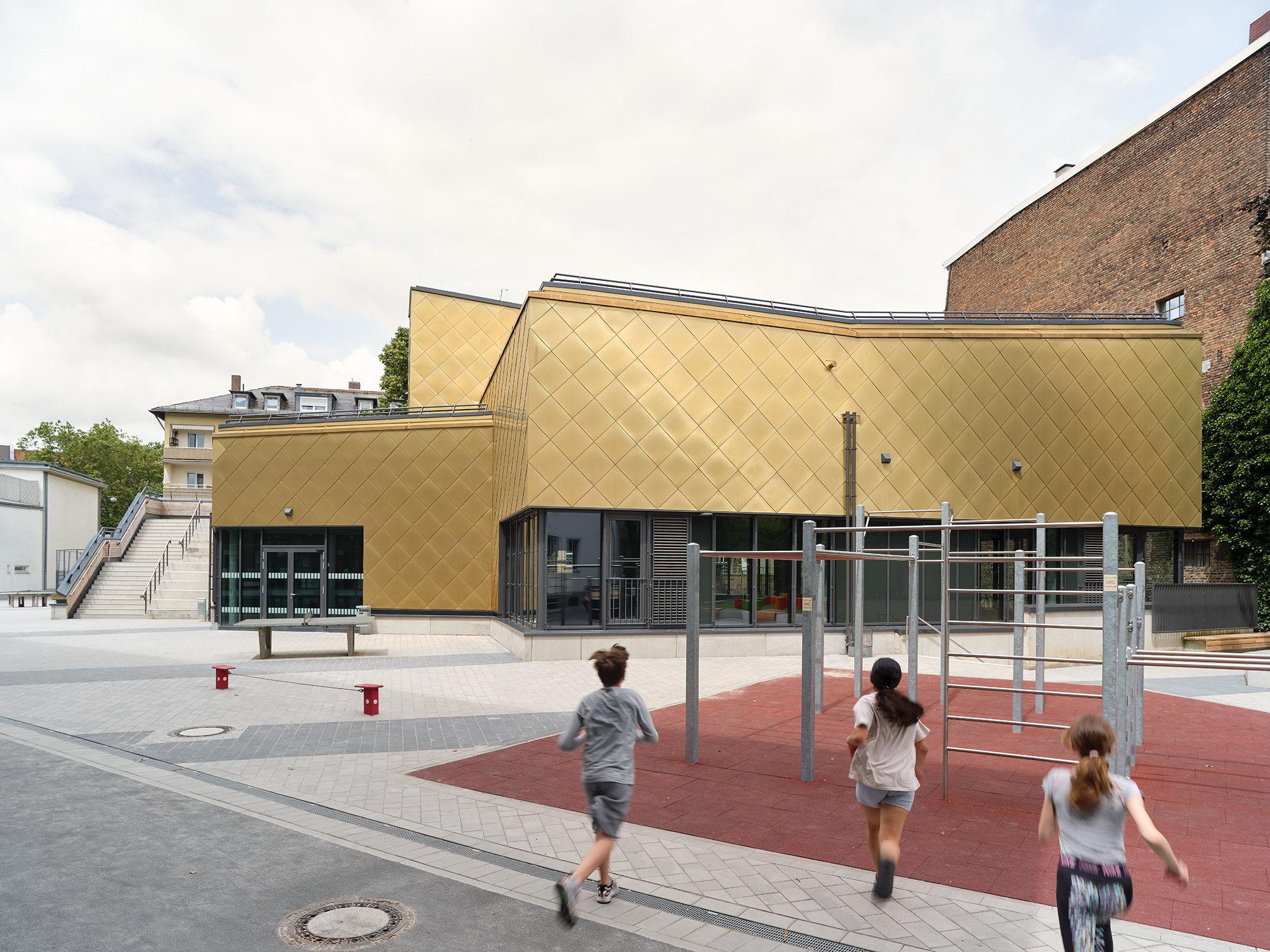 Kinder rennen über einen Spielplatz auf eine vielfach gestaffelten Sporthalle mit goldener Metalfassade zu.