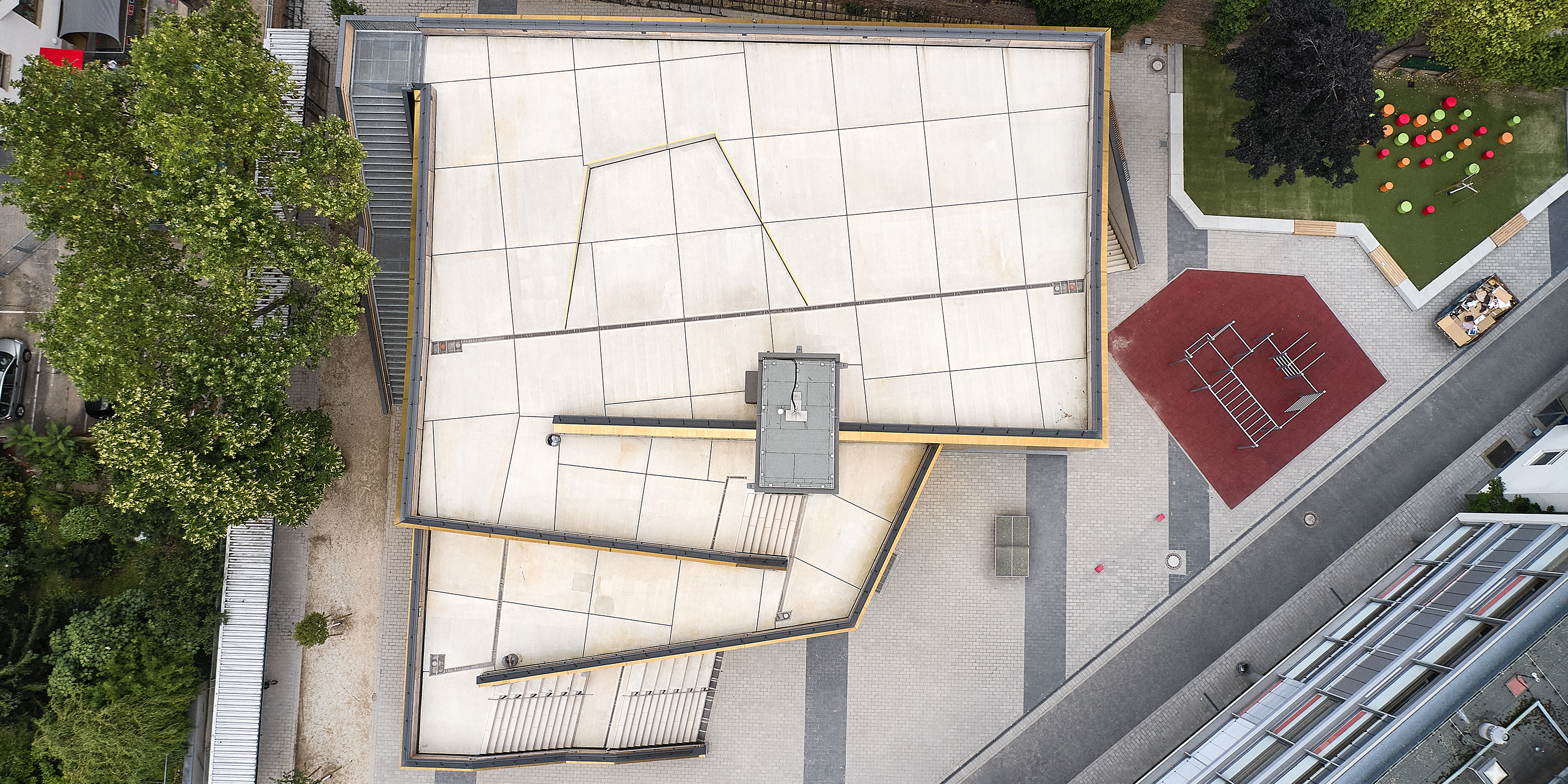 Blick von oben auf ein begehbares Dach einer mehrfach gestaffelten Sporthalle, die auf einem Schulhof steht. Ein nahe platzierter Schulbau ist im Anschnitt zu sehen.
