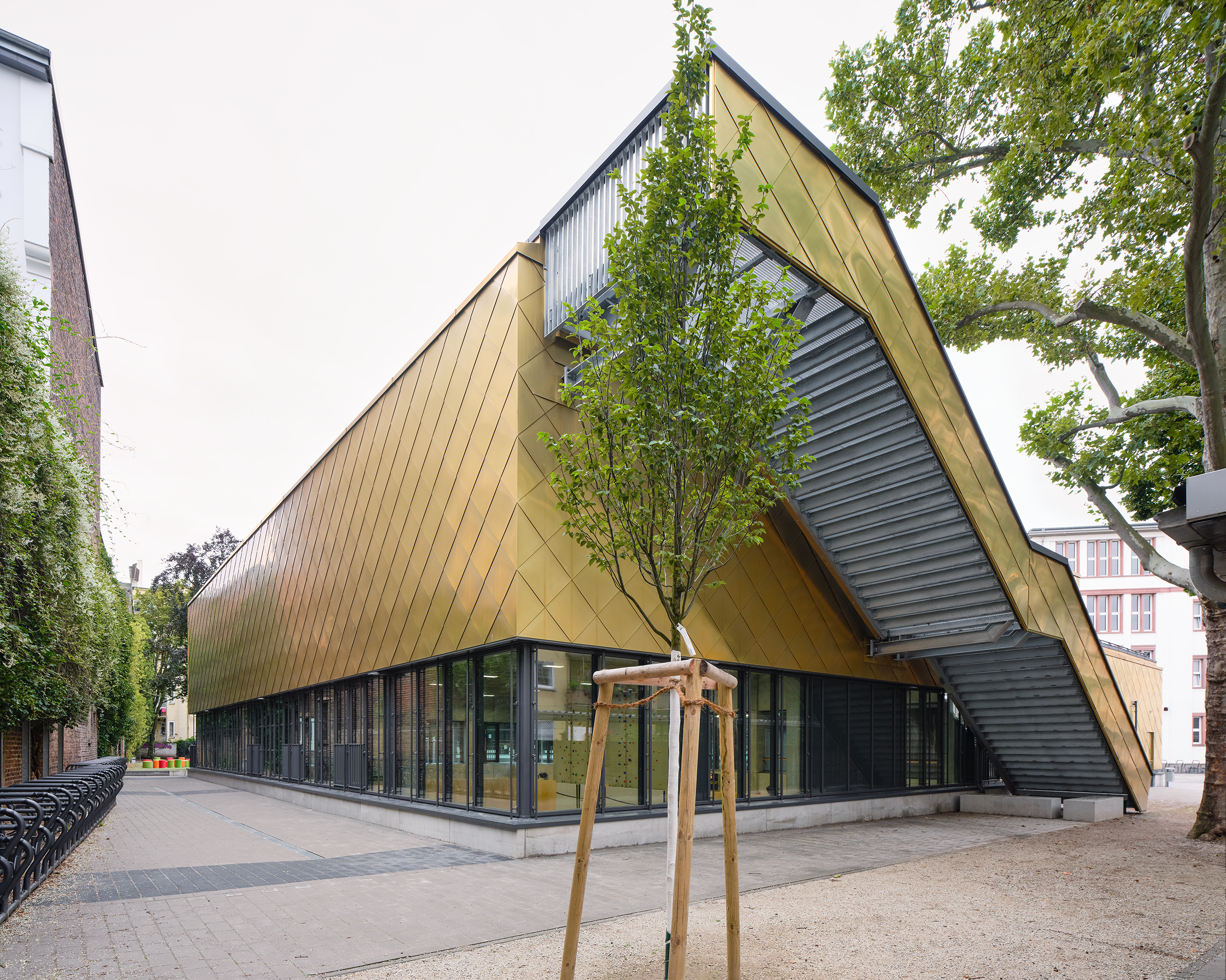 Gebäude einer Sporthalle mit Fensterband im Erdgeschoss und goldener Metallfassade im Obergeschoss. Eine Treppe führt vom Erdgeschoss auf das Dach.