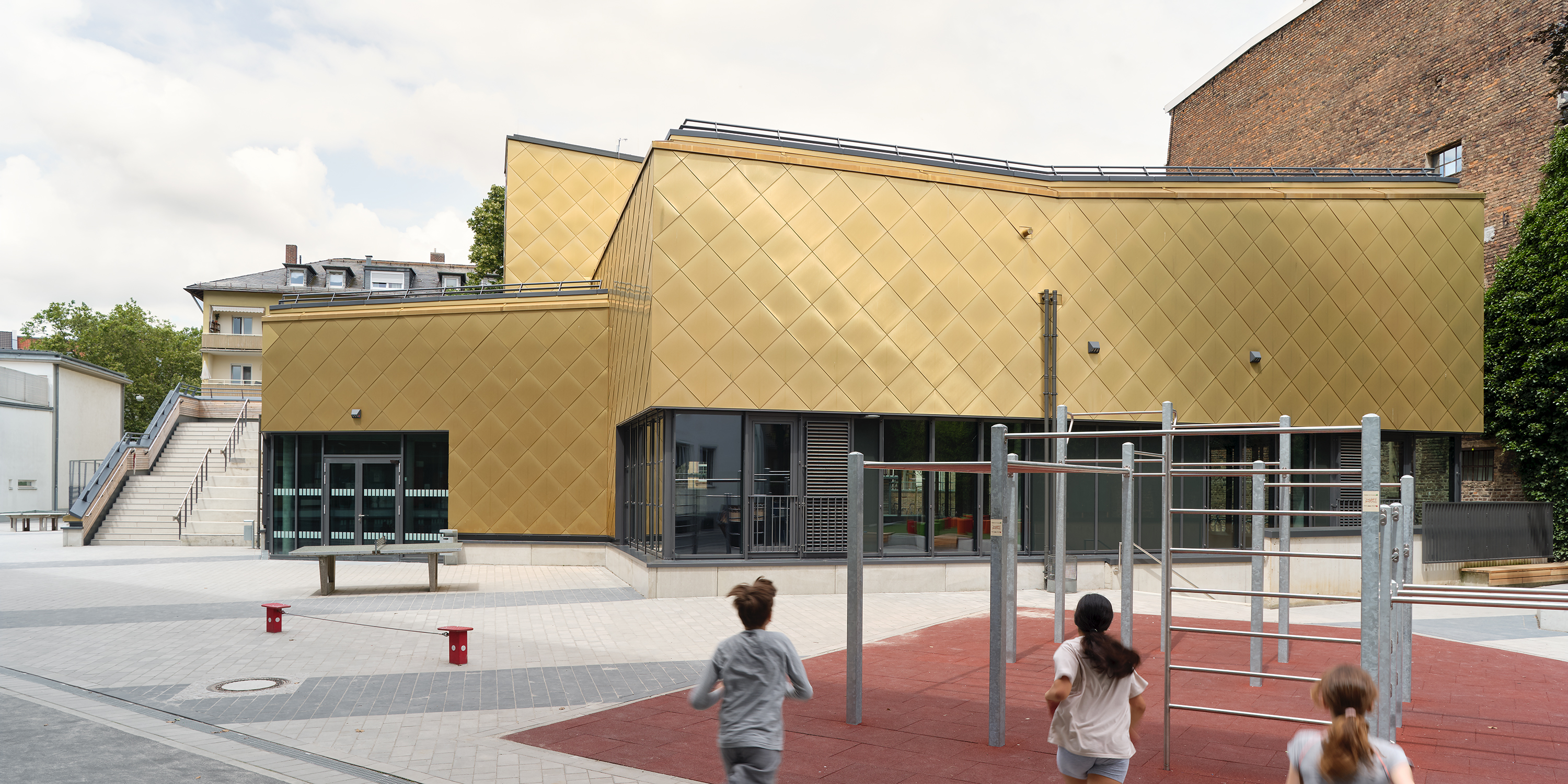 Kinder rennen über einen Spielplatz auf eine vielfach gestaffelten Sporthalle mit goldener Metalfassade zu.