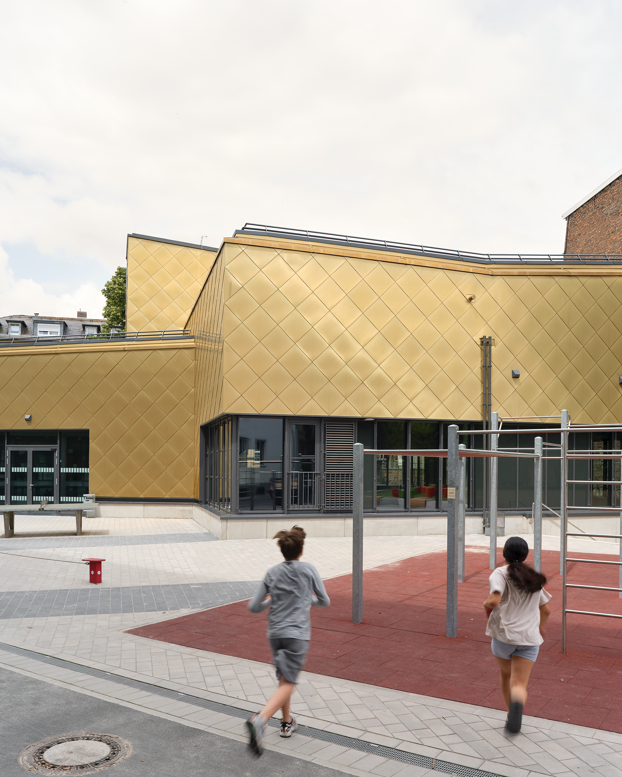 Kinder rennen über einen Spielplatz auf eine vielfach gestaffelten Sporthalle mit goldener Metalfassade zu.
