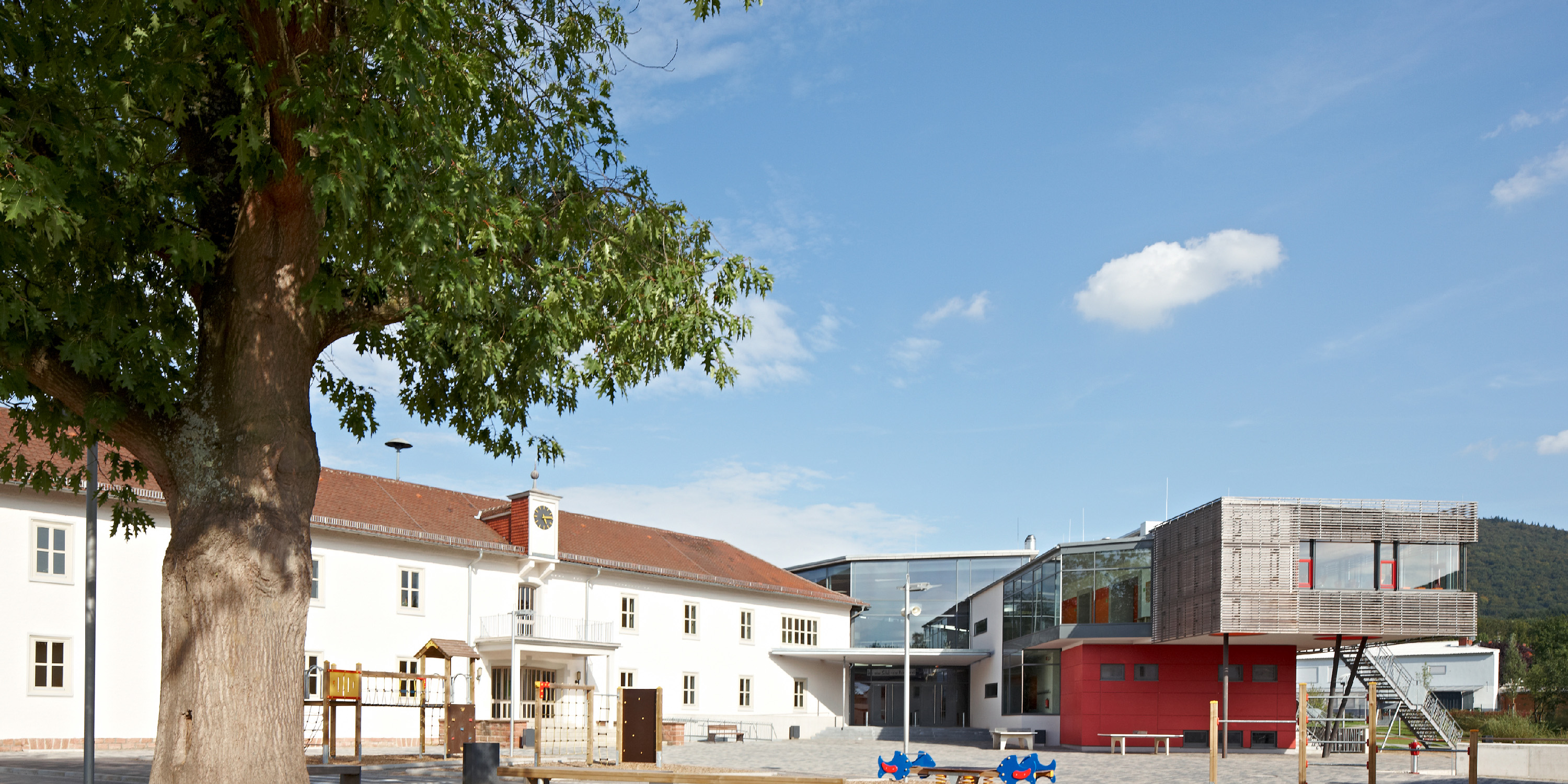 Schululbauensemble mit einem weiß verputzten Altbau und einem modernen aufgefächerten Anbau.Das Obergeschoss des Anbaus kragt aus. Dazwischen ist ein Schulhaof mit einem großen Baum und Spielgeräten