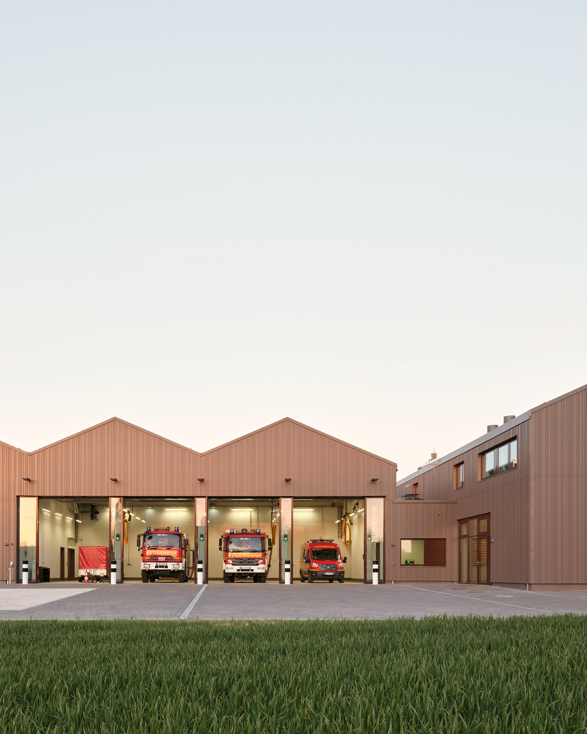 Eine Außenaufnahme eines Feuerwehrhauses mit geöffneten Toren zur Fahrzeughalle, in der vier Einsatzfahrzeuge stehen. Die Fassade ist aus rotbraunem Metall.