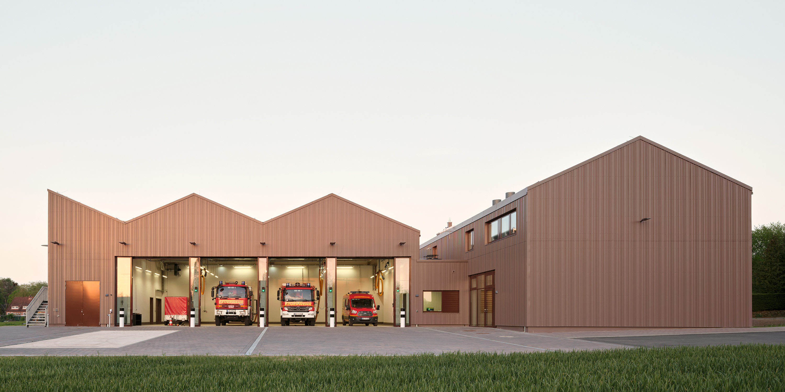Eine Außenaufnahme eines Feuerwehrhauses mit geöffneten Toren zur Fahrzeughalle, in der vier Einsatzfahrzeuge stehen. Die Fassade ist aus rotbraunem Metall.