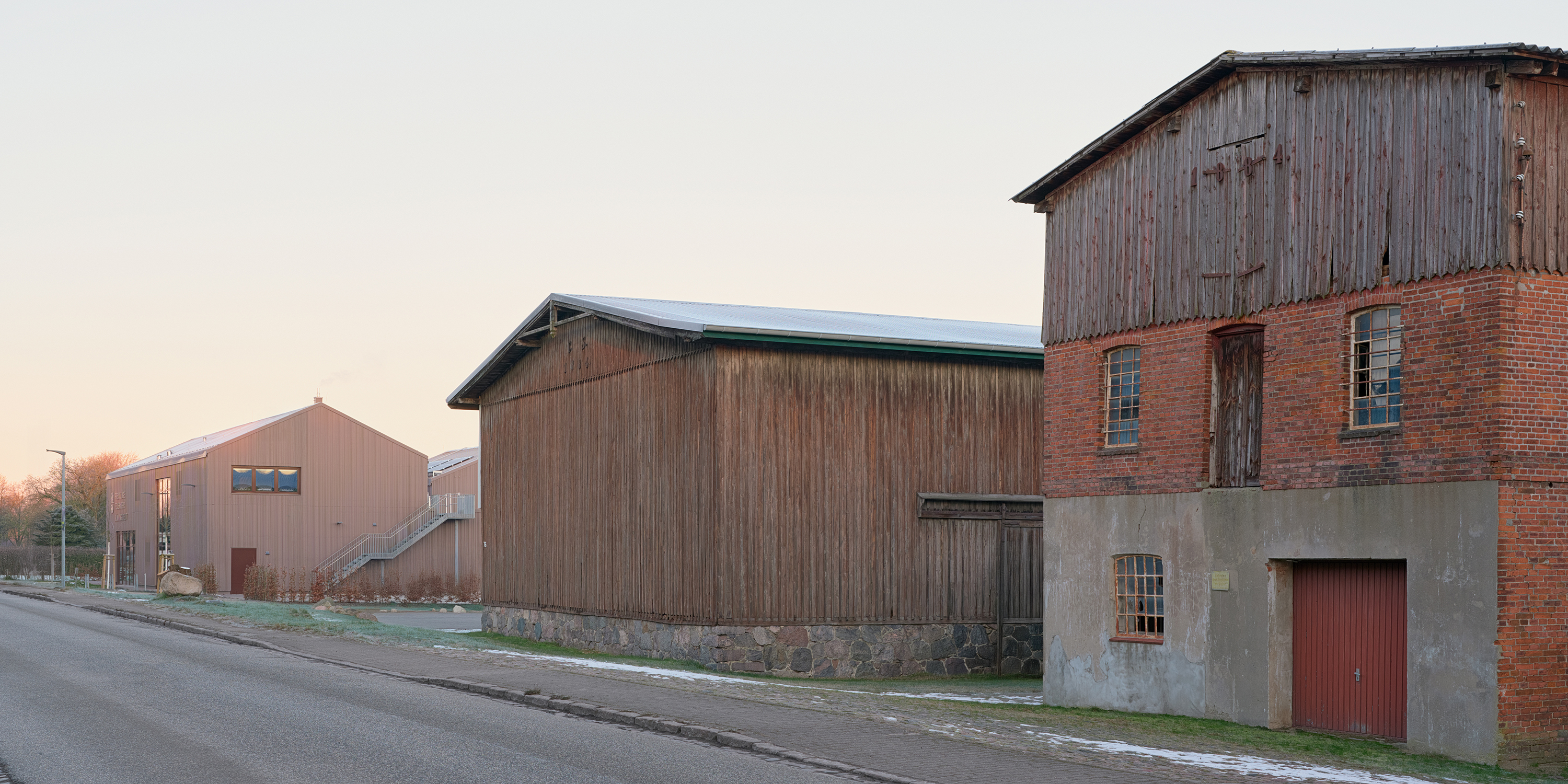 Zwei historische Scheunen mit flach geneigten Satteldächern im Vordergrund, eine Feuerwache mit rotbrauner Metallfassade und flach geneigtem Satteldach  im Hintergrund.