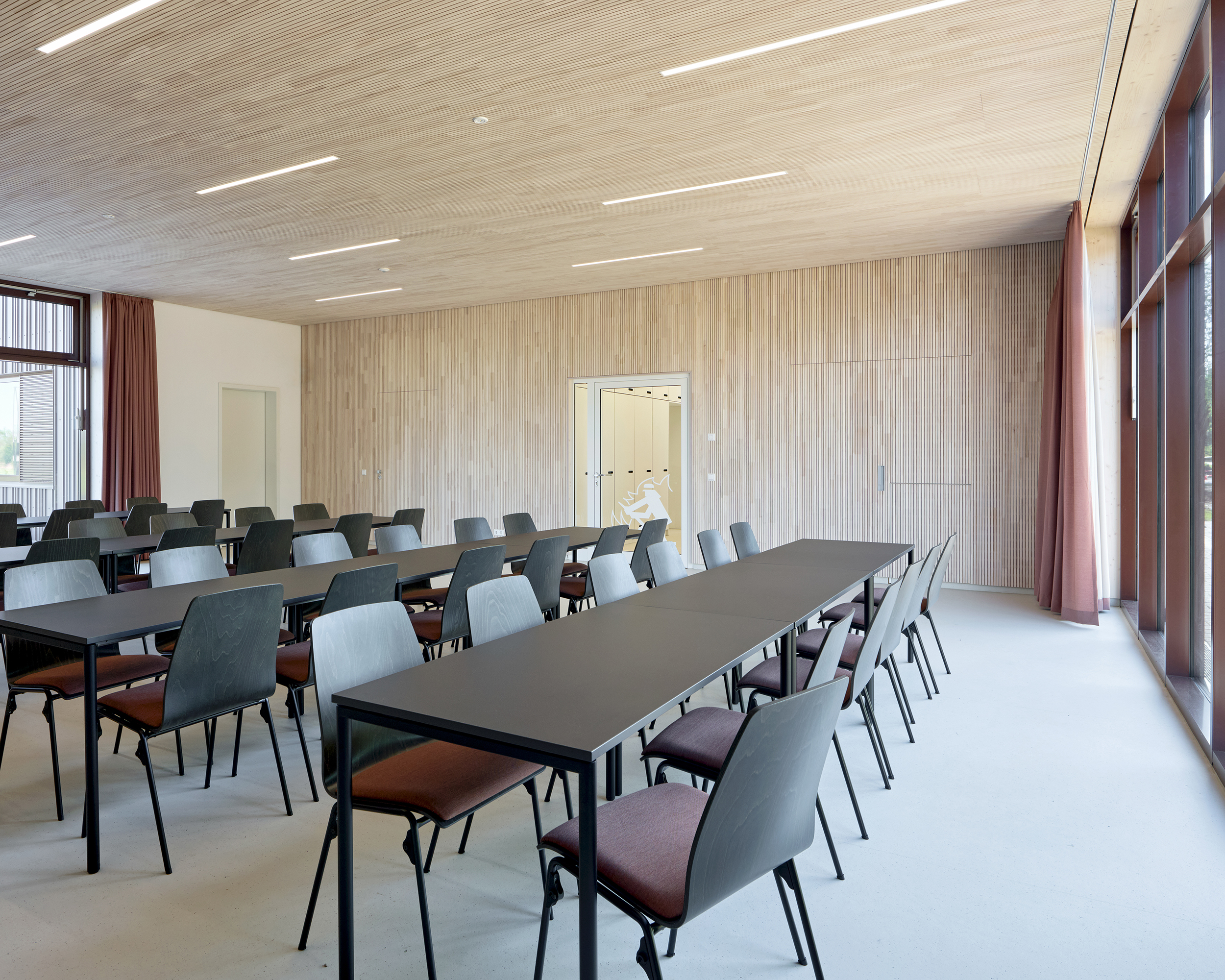Schulung- und Versammlungsraum mit langen tischen und Stühlen. Die Wände und Decke ist aus Holz. Beidseitig sind große Fenster.