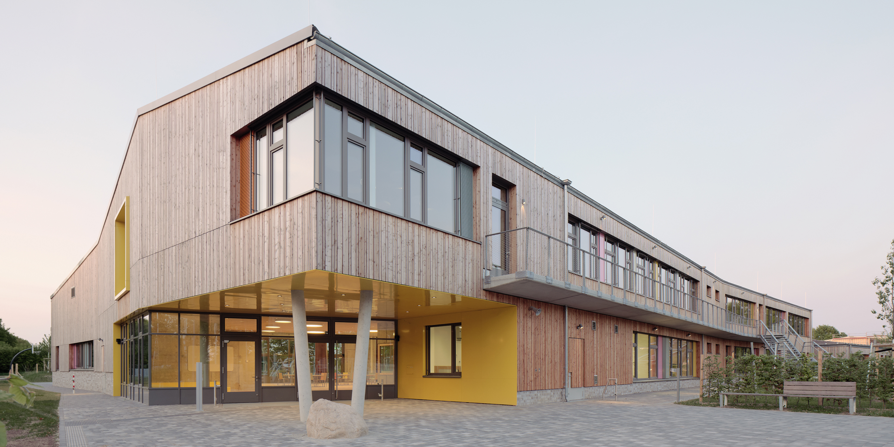 Außenaufnahme einer zweigeschossigen Schulgebäudes mit Holzfassade und und großen Fenstern. Eine Ecke des Obergeschoss kragt aus und wird von zwei schräg gestellten Stützen getragen, kräftige Farben betonen Fenster und Eingangsbereich.