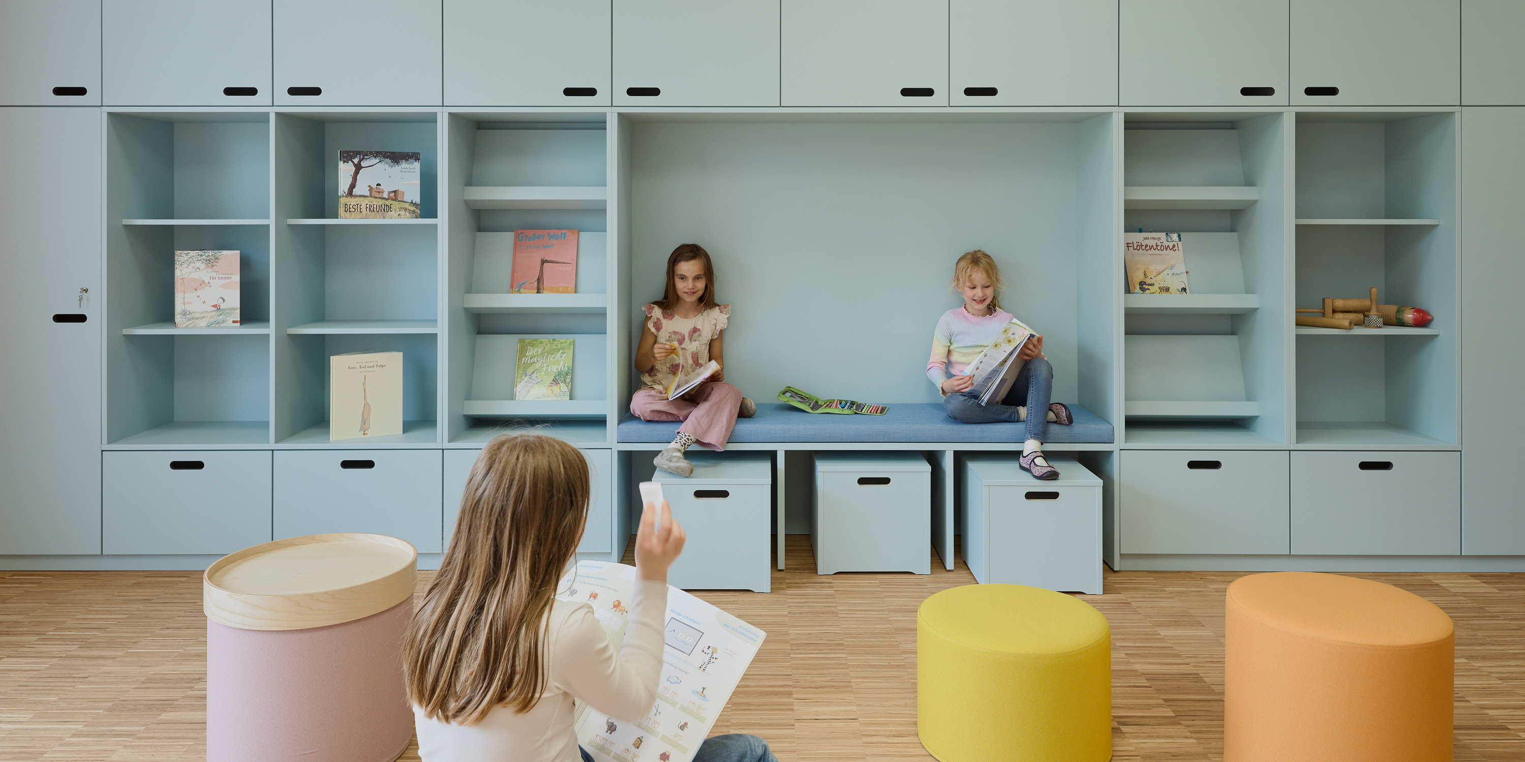 Kinder sitzen in und vor einem zartblauen Regalmöbel mit eingebauter Sitznische und lesen