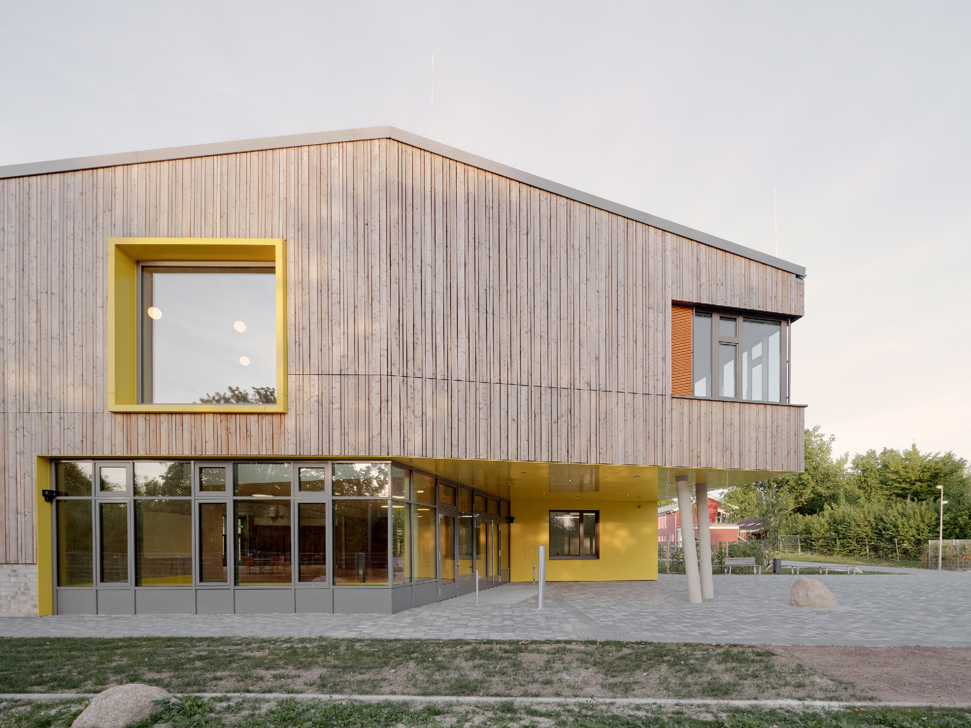 Außenaufnahme des  Eingangsbereichs eines zweigeschossigen Schulgebäudes mit flach geneigtem Satteldach, HOlzfassade und und großen Fenstern. Das Obergeschoss kragt aus, kräftige Farben betonen Fenster und Eingangsbereich.
