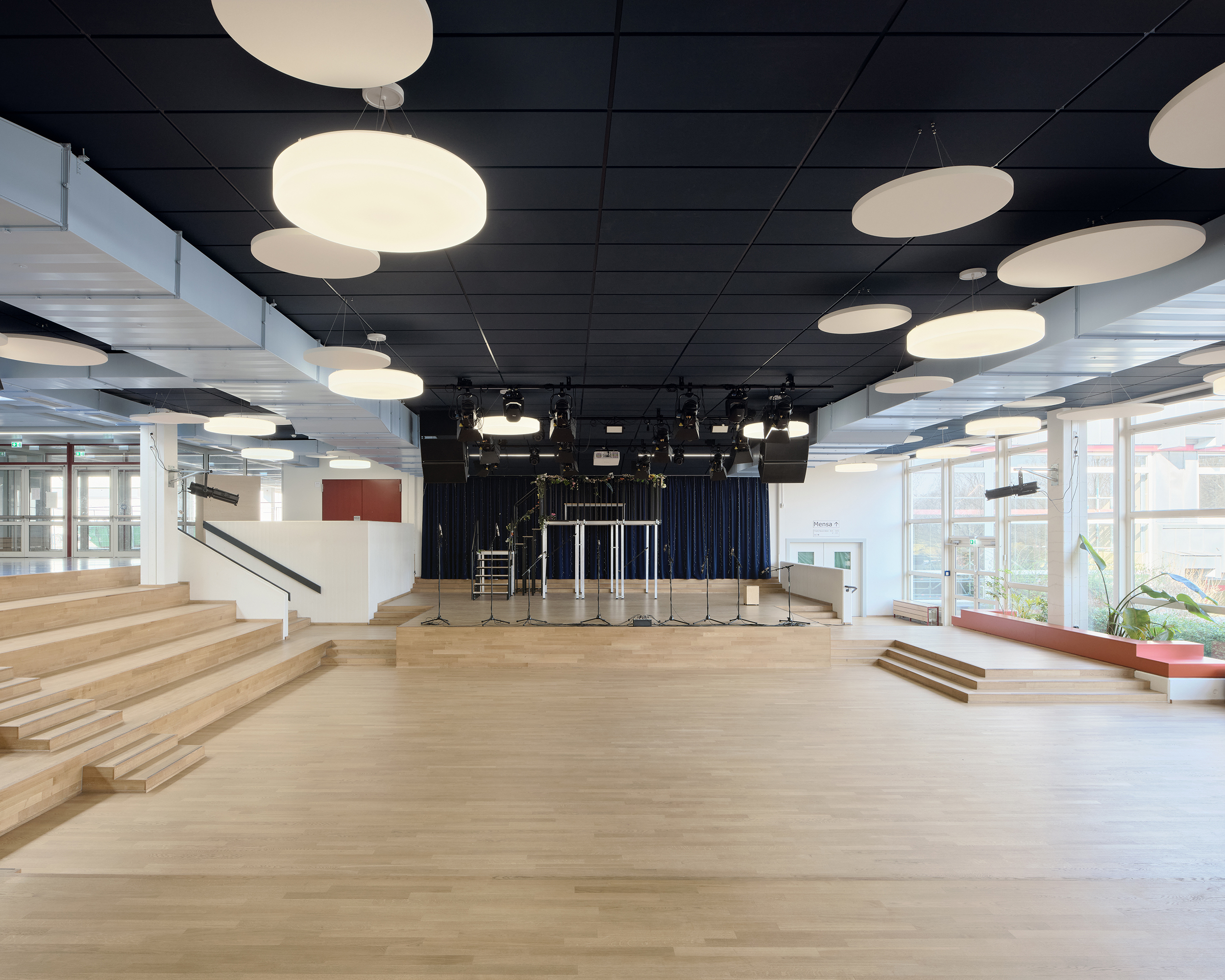 Eine Aula mit sitzstufen und Holzbodenbelag. Große, runde leuchten hängen unter der dunklen Decke.