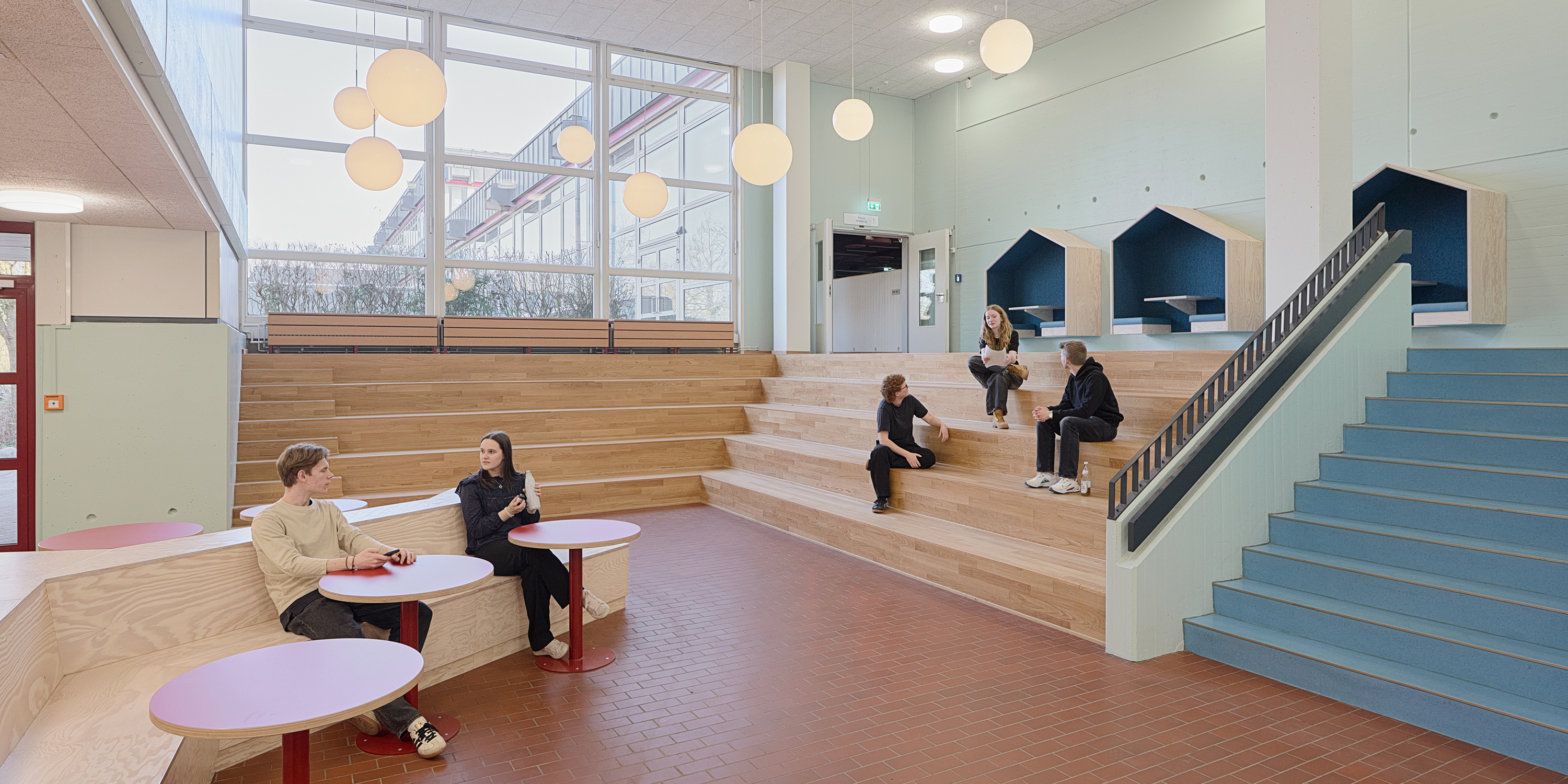 Schüler sitzen in dem Foyer einer Schule auf Sitzstufen und an fest eingebauten Möbeln mit runden Tischen.