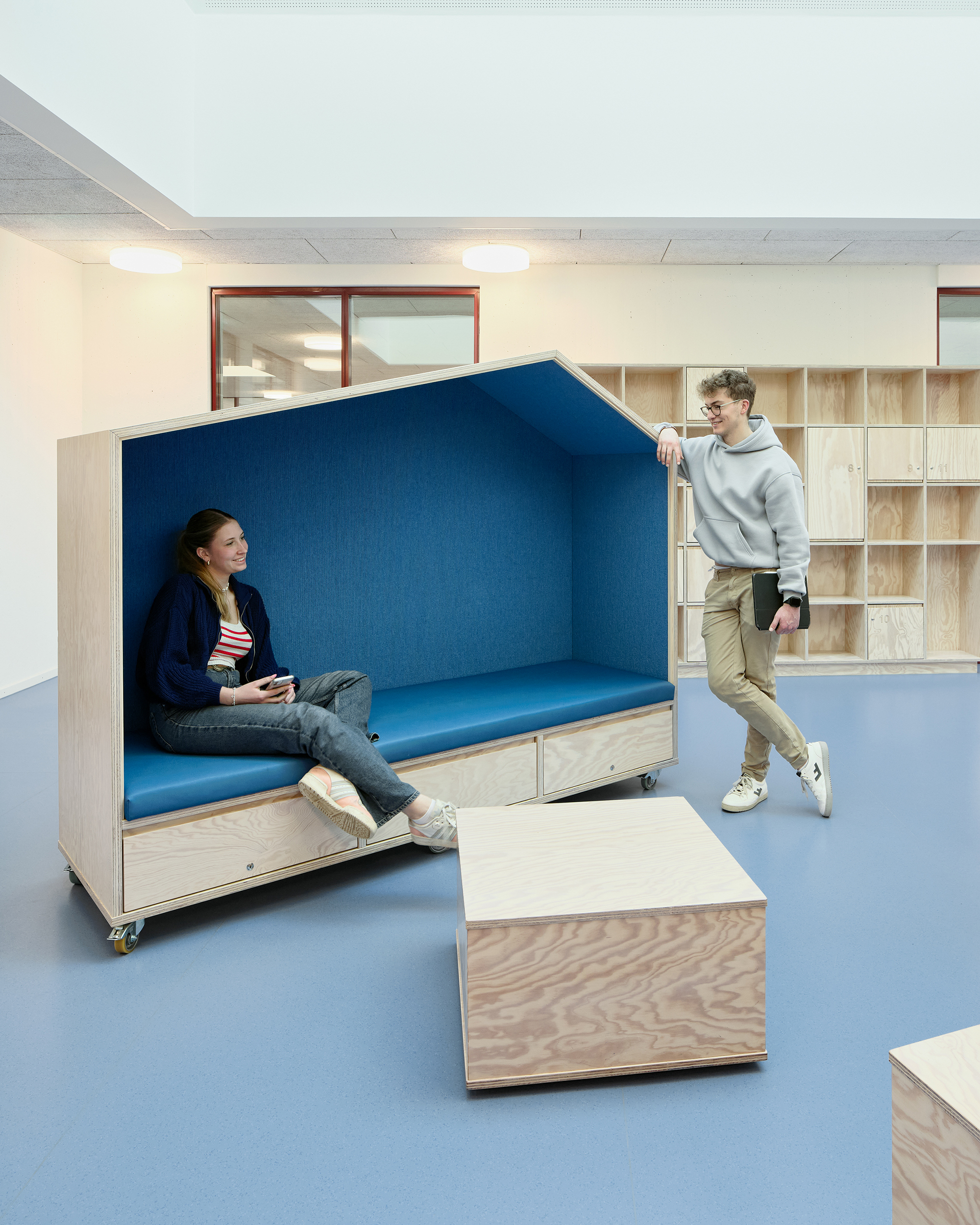 Eine Schülerin, sitzt in einem Haus Artik Sitzmöbel aus Holz. Die Innenseiten sind blau. Daneben steht ein Junge.