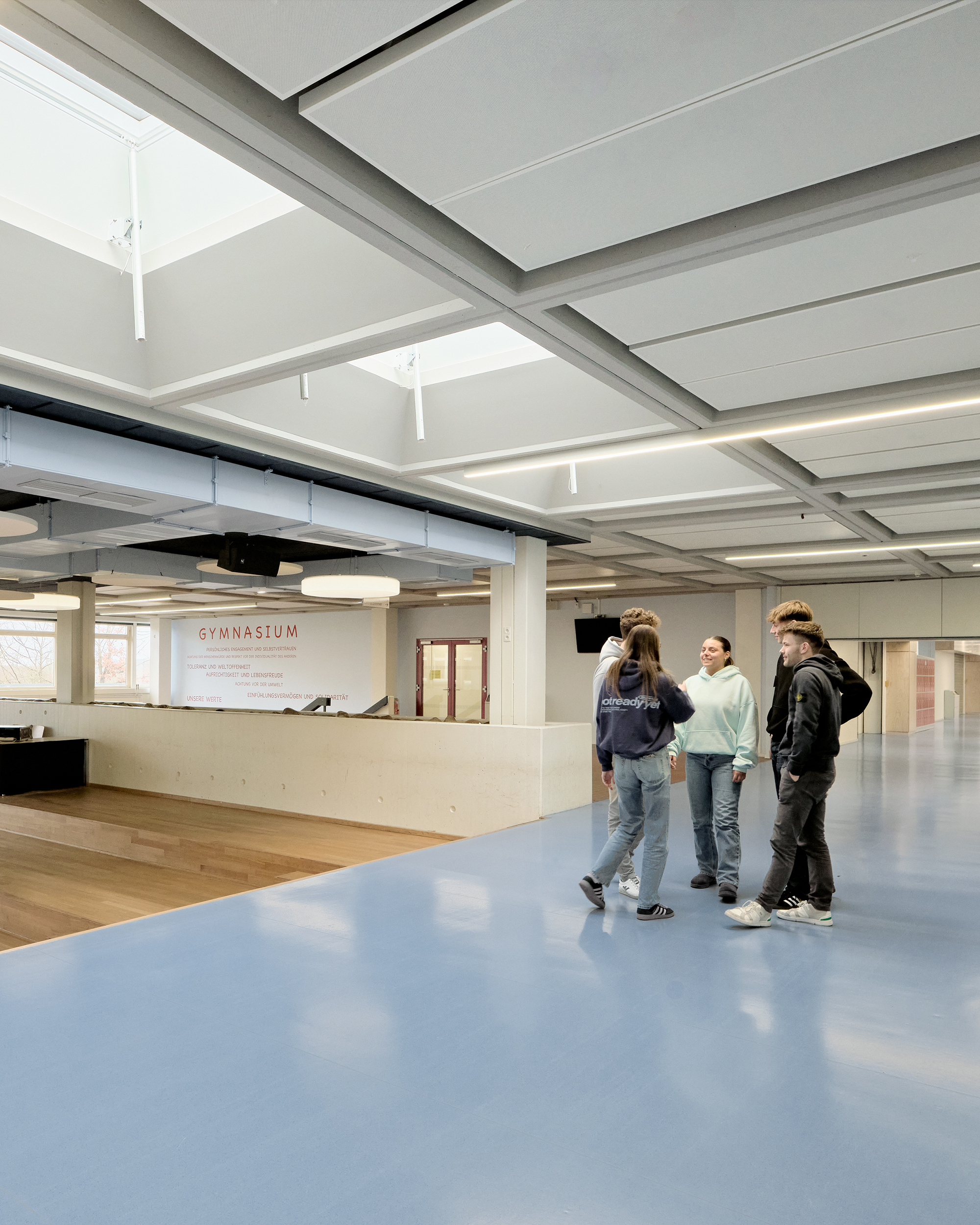 Eine Gruppe von Schülern steht in einem großzügigen Eingangsbereich einer Schule. Der Linoleum Boden ist hellblau, die Decke ist gerastet.