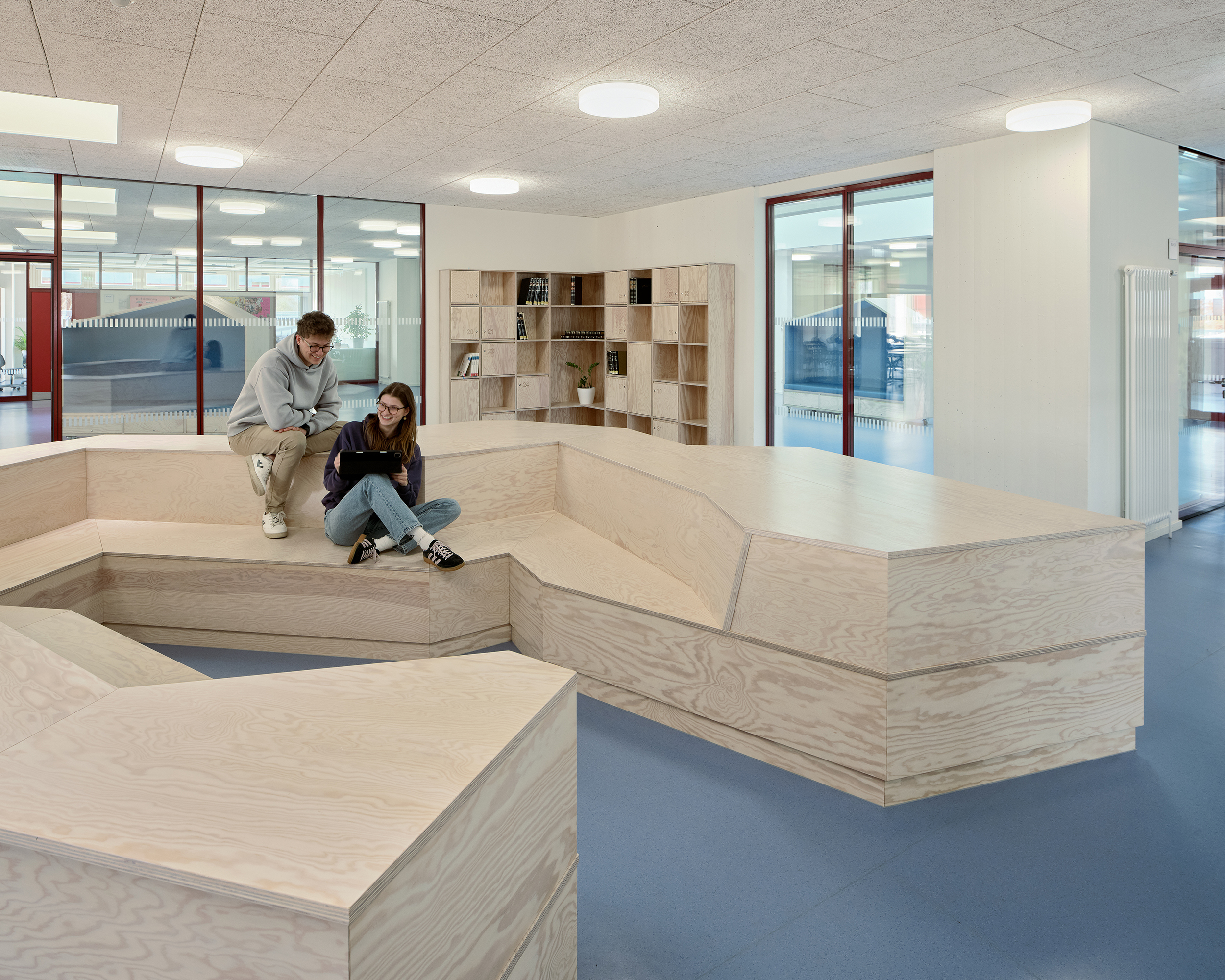 Zwei Schüler sitzen auf einem mehrfach geknickten Sitzmöbel aus Holz. Für Inoculum Boden ist hellblau. Im Hintergrund blickt man in die angrenzenden Räume.