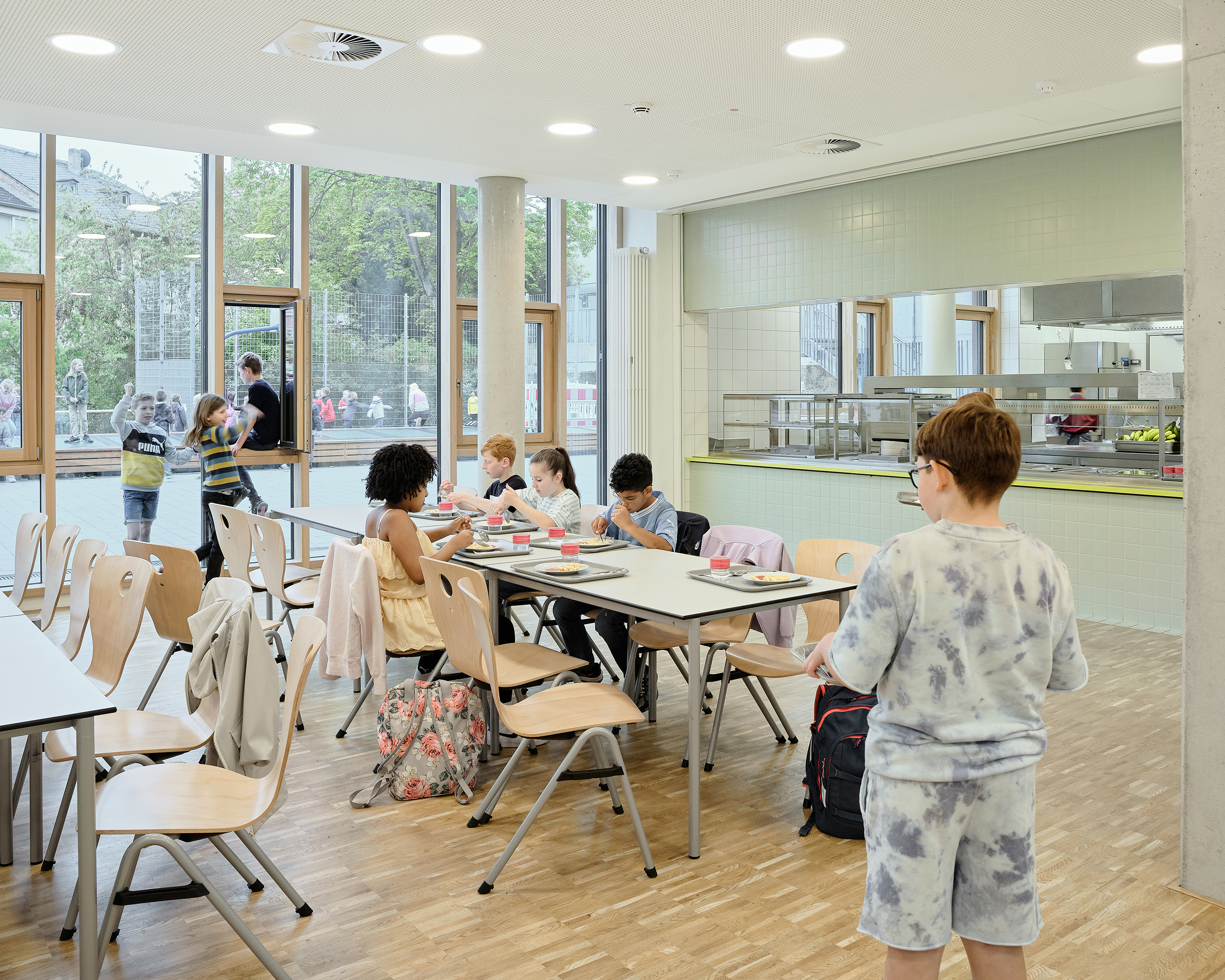 Kinder sitzen und stehen in einer Mensa an langen Tischen. Eine große Glasfassade richtet sich zumn Schulhof