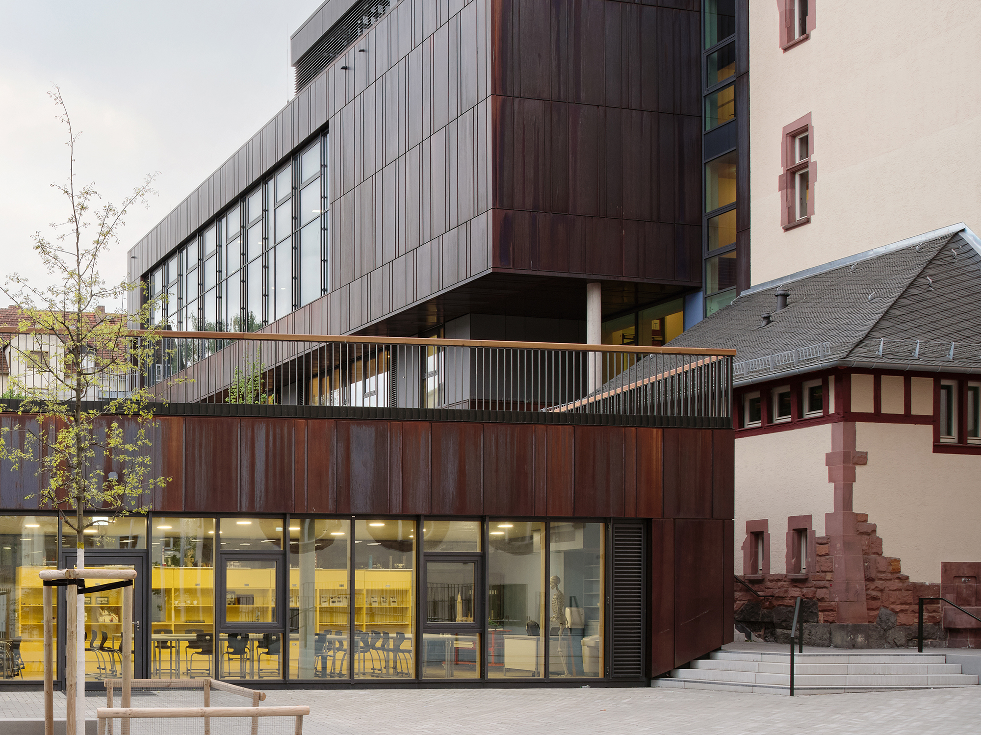 Ein Schulneubau mit dunkler Metallfassade und Schulhof. Aus einer großen Galsfassade scheint warmes Licht.