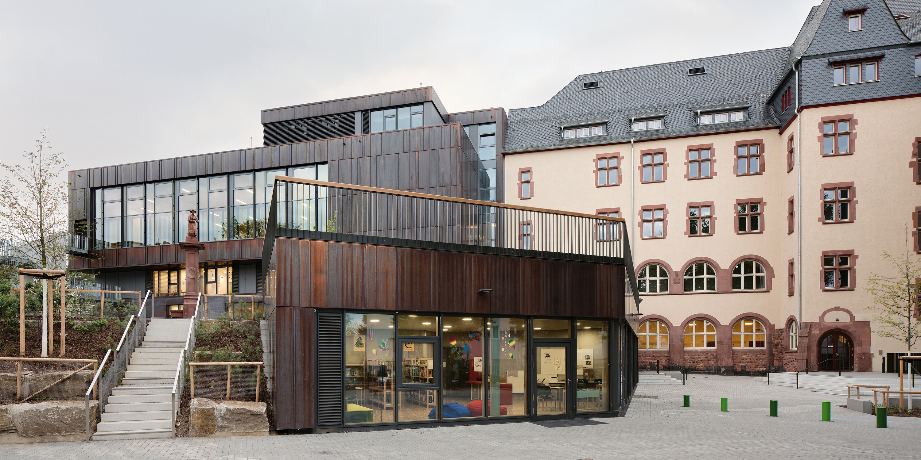 Ein Schulneubau mit dunkler Metallfassade und Schulhof. Eine Treppe führt auf einen höher gelegenen Schulhof
