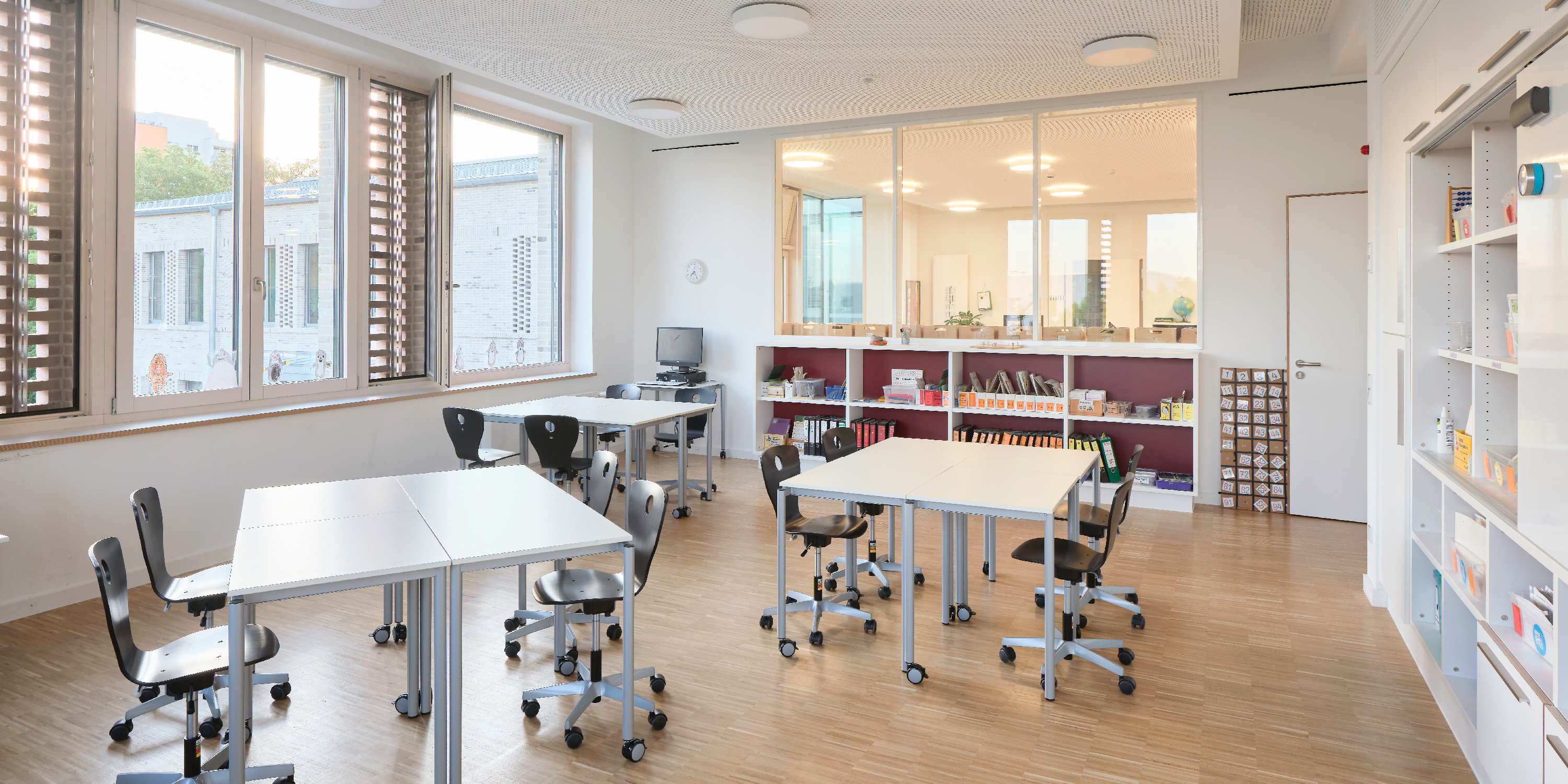 Unterrichtsraum einer Schule mit locker aufgestellten Gruppentischen, Regal und Sichtbeziehung zur gemeinsamen Mitte.